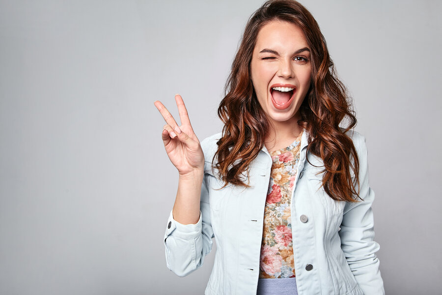 Portrait of young stylish laughing girl model in colorful casual summer clothes with natural makeup isolated on gray background. Looking at camera and showing peace sign