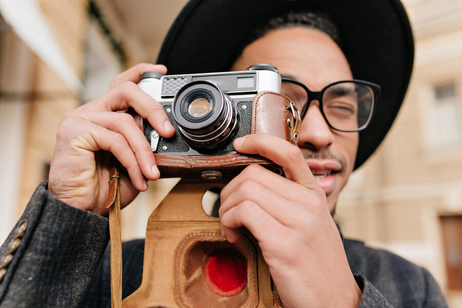 Close-up portrait of handsome black man in elegant glasses making photos with camera. Concentrated african photographer working outdoor.