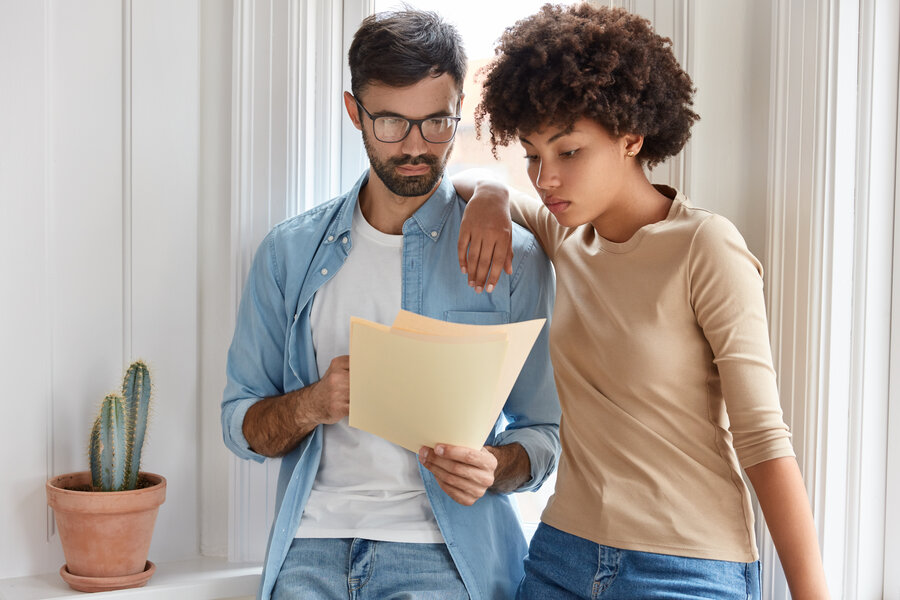 Image of successful young coworkers focused in documents recieved from bank, ready to buy renting apartment, ready to have deal with realtor, stand closely nearwindow, study contract before singing